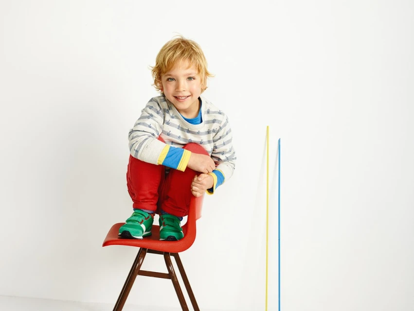 A young child with blond hair poses on a red chair in a studio, wearing a striped shirt, red pants, and green sneakers. 
