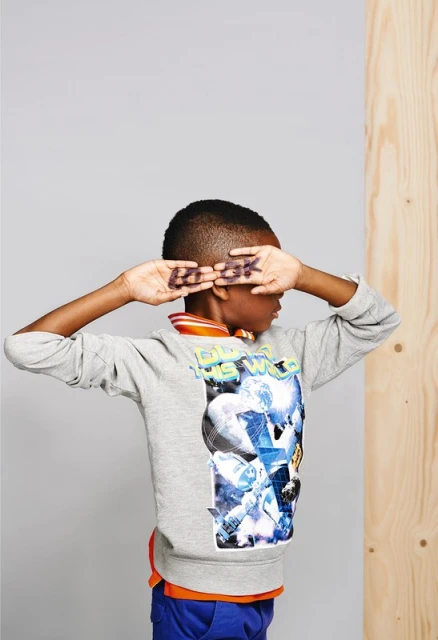 Young boy poses in a gray sweatshirt and blue pants, hands over his eyes, with the word 'Rock' written on his hands in studio shot.