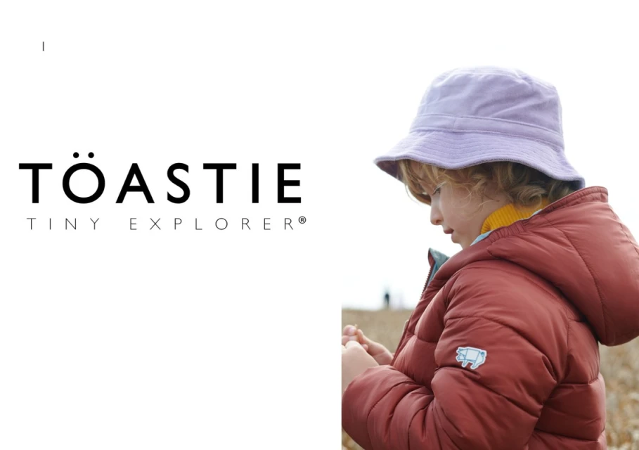 Child in rust-colored puffer jacket and lilac bucket hat outdoors. Warm color palette, natural light, fashion-forward kidswear.