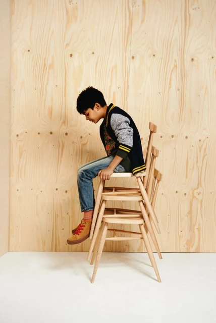 Young boy sits atop stacked wooden chairs in a minimalist studio setting, wearing a casual outfit and brown shoes, with a wood panel backdrop.