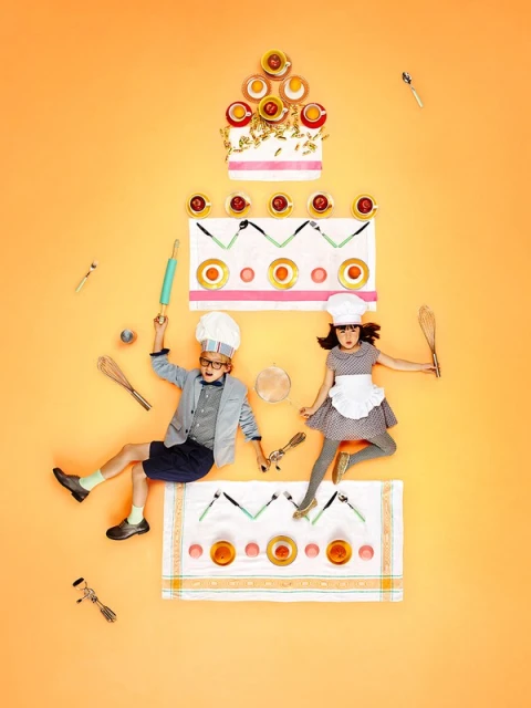 Overhead shot of two children wearing chef hats lying near kitchen utensils arranged like a cake on an orange background.