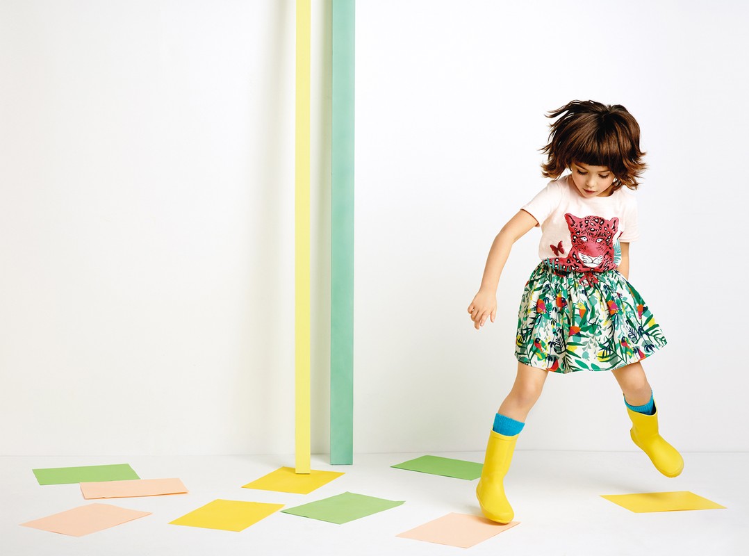 A girl with brunette hair jumps in the air wearing a t-shirt with an animal print and a skirt with a leaf pattern, along with yellow boots.