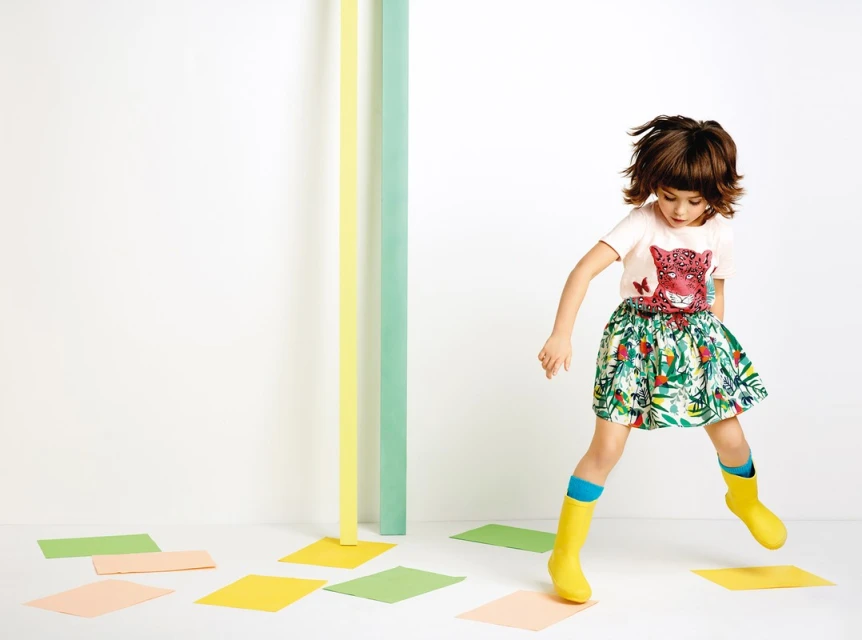 A girl with brunette hair jumps in the air wearing a t-shirt with an animal print and a skirt with a leaf pattern, along with yellow boots.