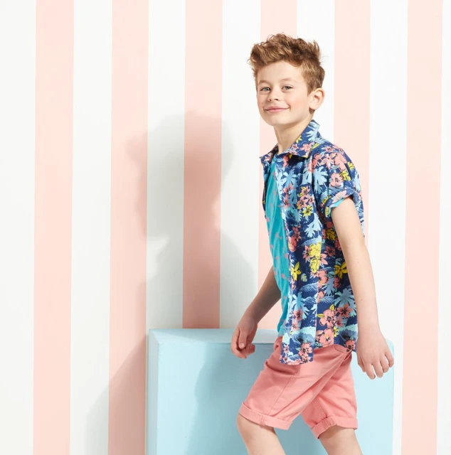 Boy in a floral shirt and coral shorts walking in front of a striped wall and blue block, looking at the camera with a smile.