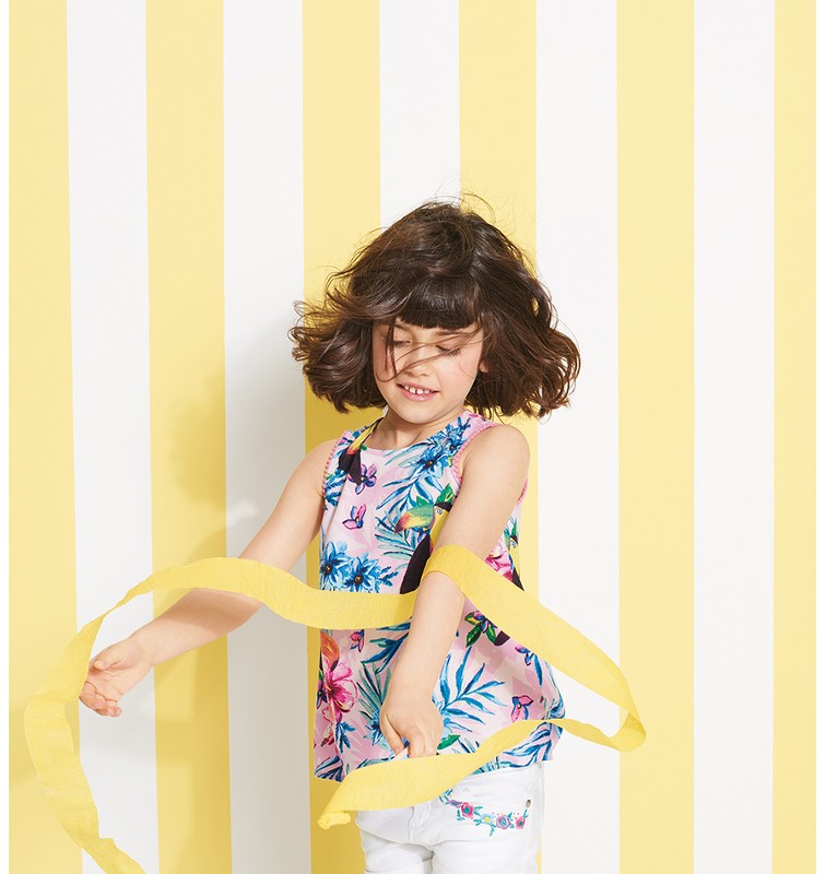 A young girl in a floral top plays with a yellow ribbon against a striped yellow and white background, captured in a studio setting.