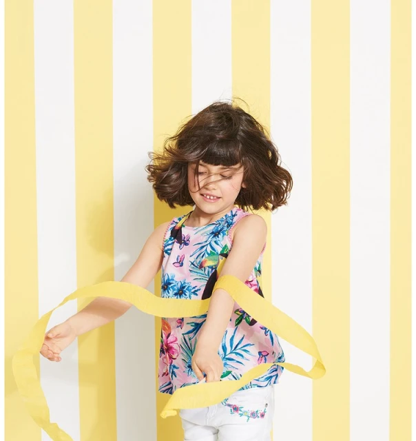 A young girl in a floral top plays with a yellow ribbon against a striped yellow and white background, captured in a studio setting.