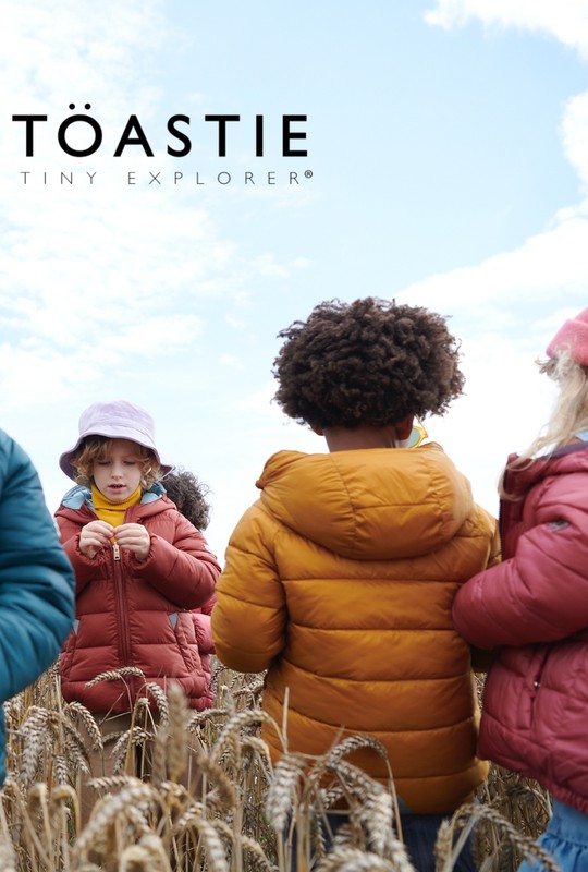Group of children in puffer jackets in a wheat field with overcast sky, Toastie Tiny Explorer branding visible, shot at eye-level.