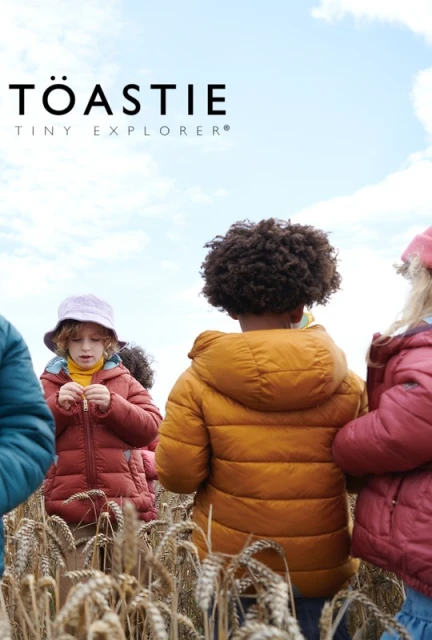 Group of children in puffer jackets in a wheat field with overcast sky, Toastie Tiny Explorer branding visible, shot at eye-level.