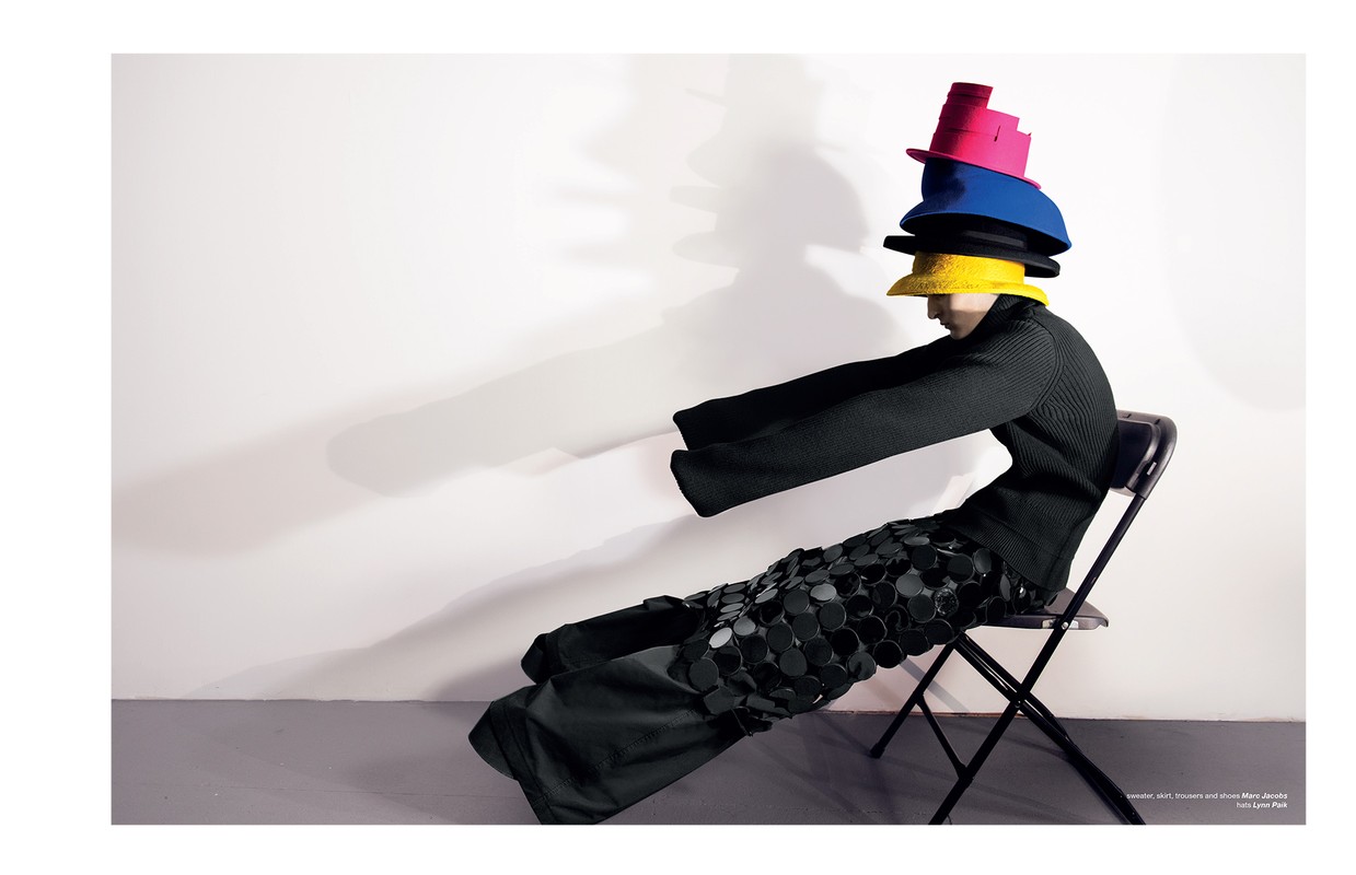 Fashion editorial image featuring a seated person wearing a black outfit and a stack of colorful hats against a white backdrop.
