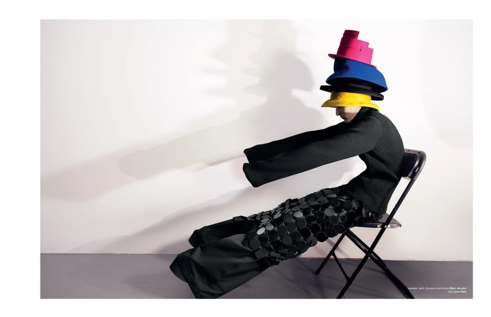 Fashion editorial image featuring a seated person wearing a black outfit and a stack of colorful hats against a white backdrop.