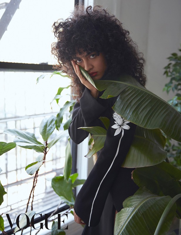 Indoor portrait of a curly-haired woman in a black top among lush houseplants near a window in natural light. Vogue logo visible.