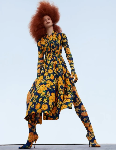 A woman with an afro poses in a blue, yellow, and green floral dress, tights, and high heels against a light blue background, angled wide shot.