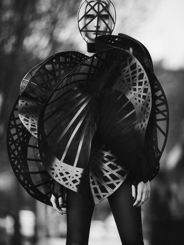 Monochrome fashion shot of a woman wearing a unique headpiece and large, geometrically patterned sculptural dress in an outdoor setting.