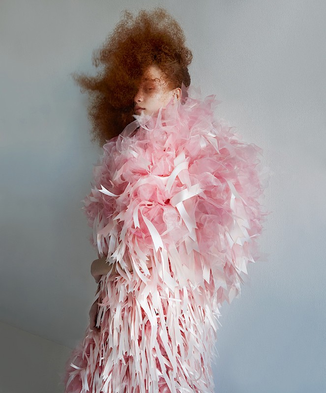 Adult female poses in a pink gown with long ribbon-like accents. The shot is a studio portrait with a neutral backdrop, fashion-focused.
