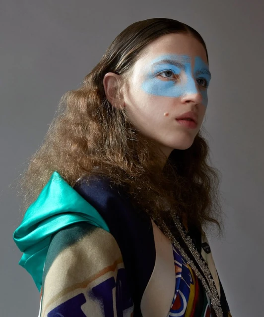 Headshot of a young woman with wavy brown hair, turquoise face paint, and a multi-patterned top, posing against a grey background.