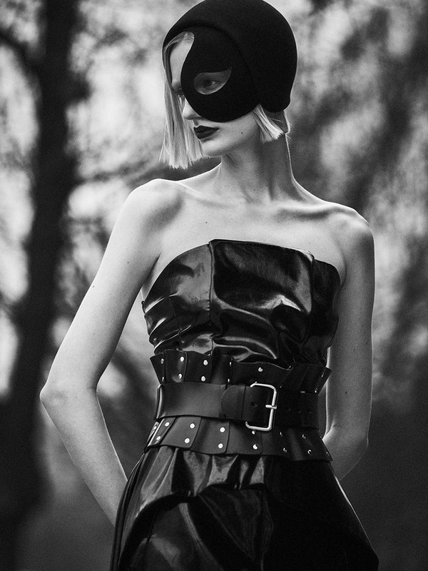 A woman with short blond hair poses in a black strapless dress and a black mask in an outdoor setting with a blurred background.