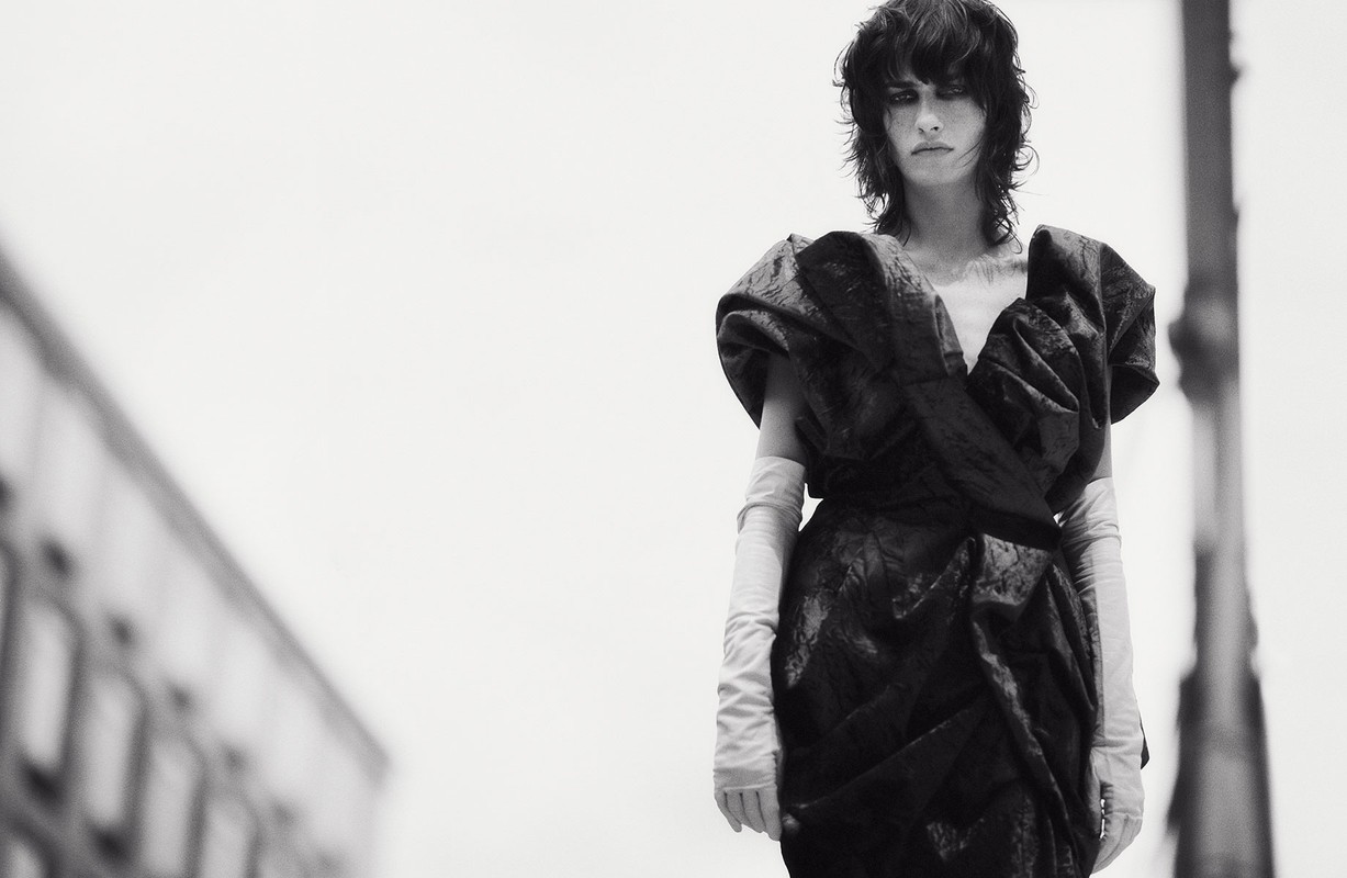Monochrome portrait of a woman in an elegant black dress with sculpted bodice, paired with long gloves, in an outdoor urban environment.