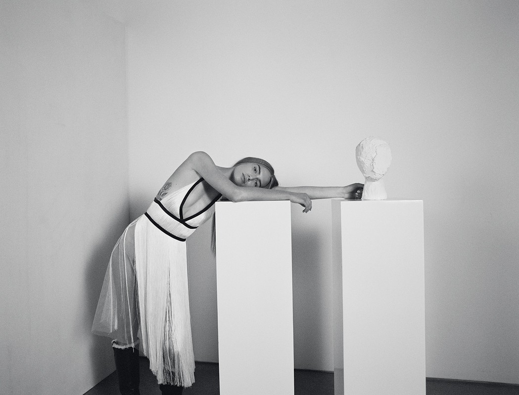 Monochrome portrait of a woman in a white dress leaning against white pedestals with a plaster head in a minimalist indoor setting.