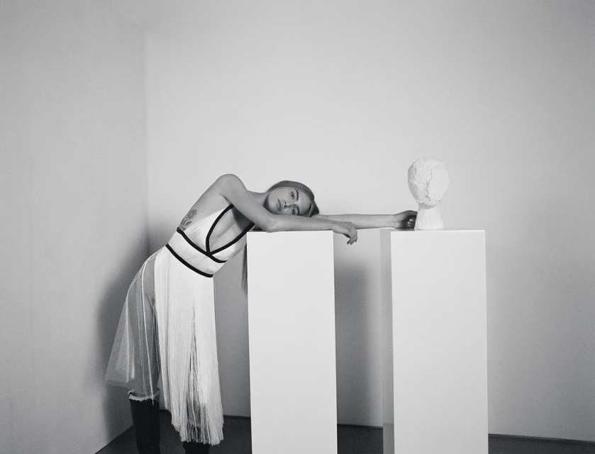 Monochrome portrait of a woman in a white dress leaning against white pedestals with a plaster head in a minimalist indoor setting.