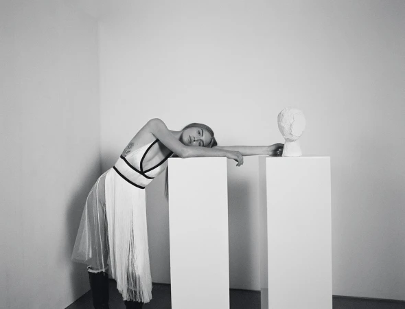 Monochrome portrait of a woman in a white dress leaning against white pedestals with a plaster head in a minimalist indoor setting.