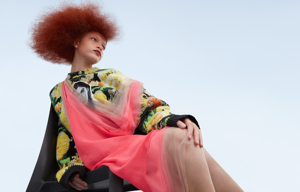 Young woman with afro hairstyle poses in a floral print sweater dress with coral fabric, seated on a black chair against a light blue sky.