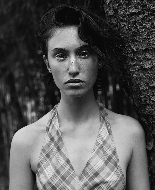 Portrait of a young woman with shoulder-length brown hair, wearing a plaid top, standing next to a tree, looking directly at the camera.