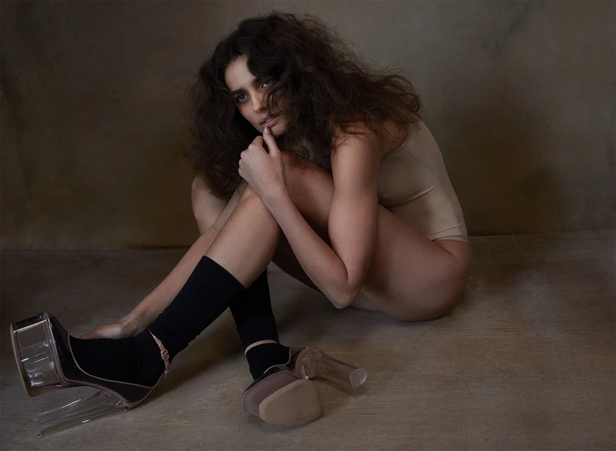 A young woman with curly hair, wearing a beige bodysuit and black socks, sits in heels indoors against a neutral background.