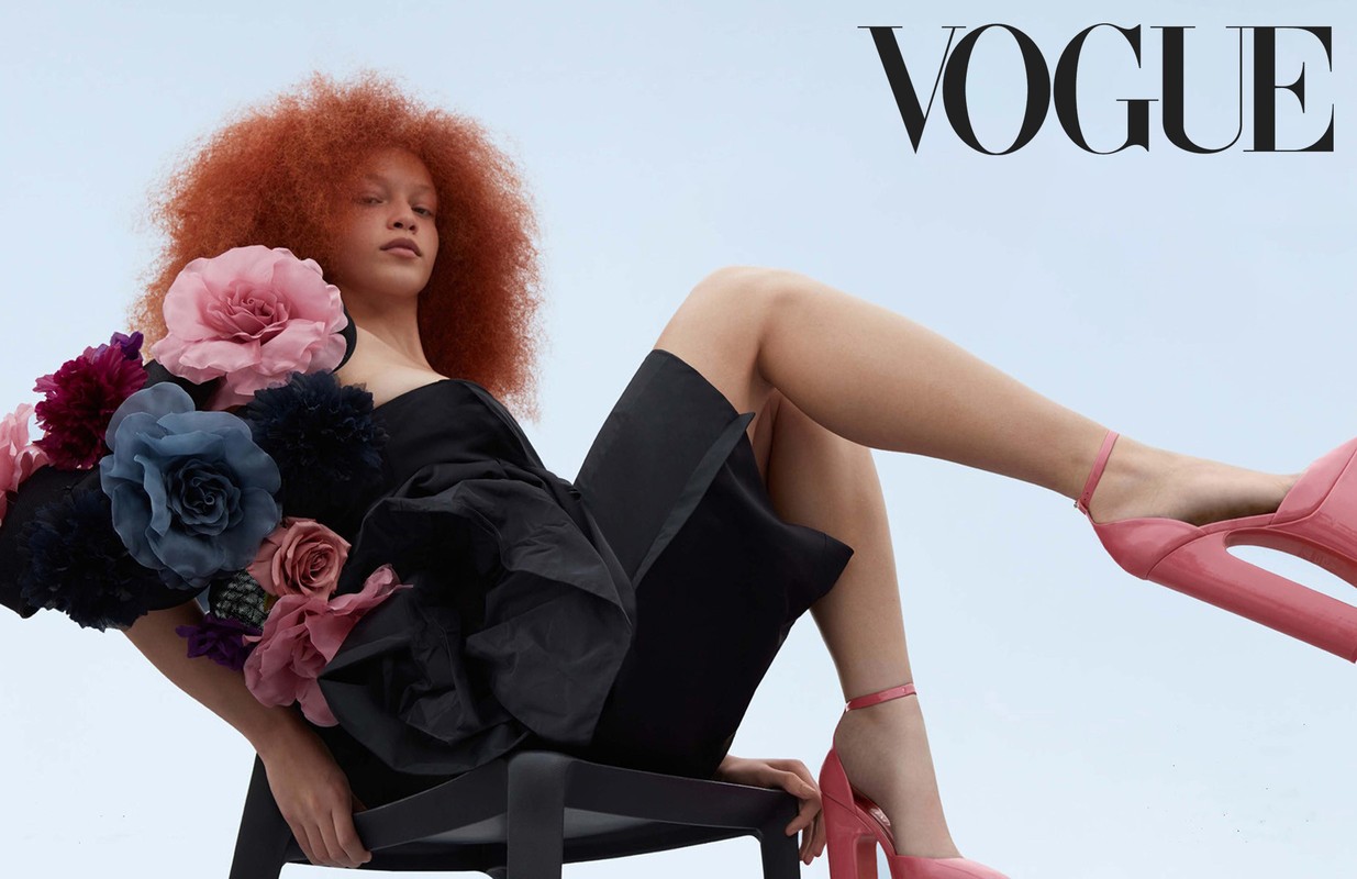 Fashion editorial image: a redhead model poses in a sculptural black dress adorned with oversized fabric flowers and pink heels against a gradient sky.
