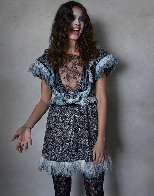 A young adult woman in a grey patterned minidress with lace trim poses in front of a neutral grey background with a big smile.