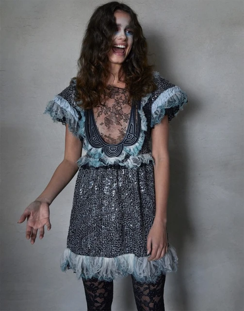 A young adult woman in a grey patterned minidress with lace trim poses in front of a neutral grey background with a big smile.