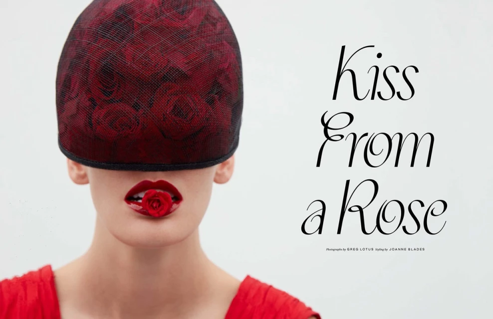 Close-up fashion shot of a woman with a red rose in her mouth and a rose-patterned cap obscuring her eyes against a bright background.