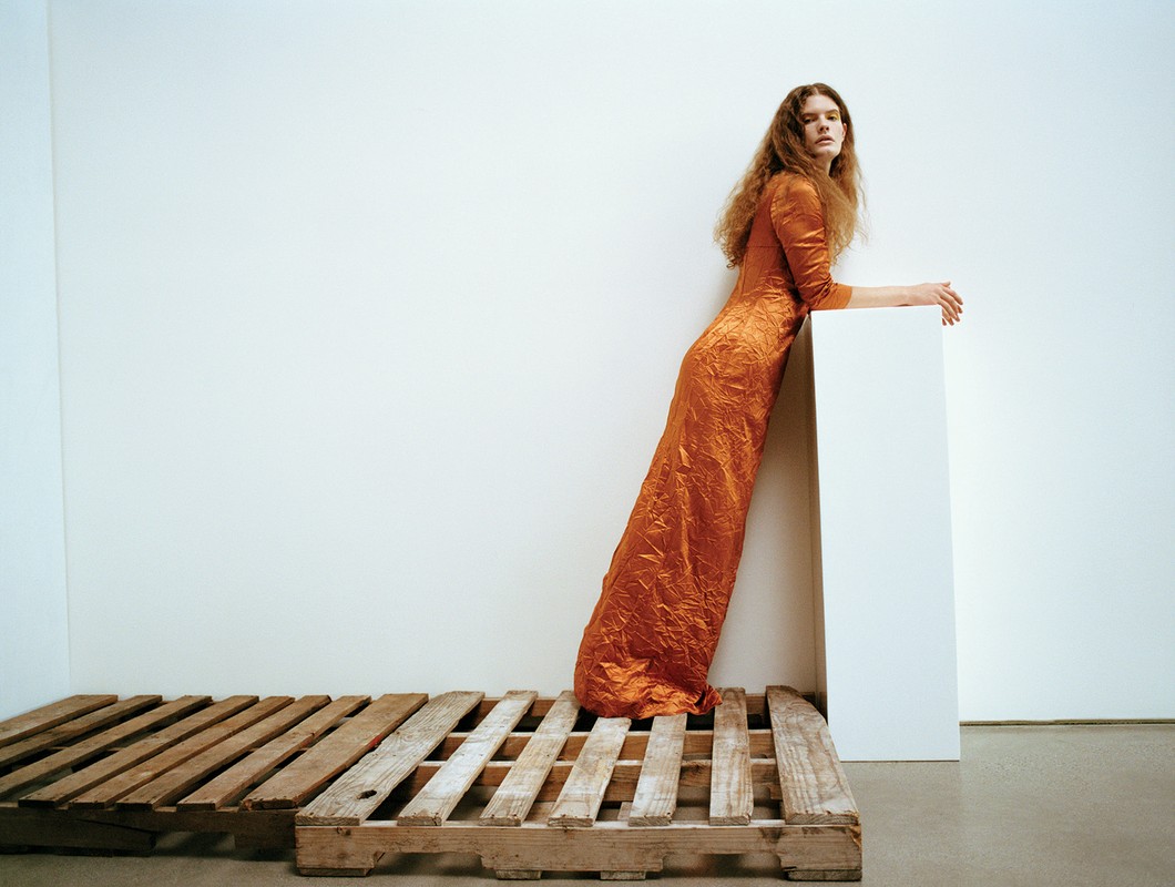 A woman in an orange dress leans against a white block, posing on a wooden pallet in a minimalist indoor setting with a white backdrop.