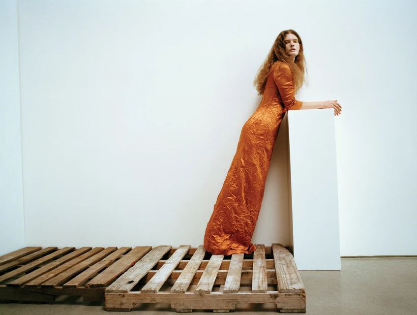 A woman in an orange dress leans against a white block, posing on a wooden pallet in a minimalist indoor setting with a white backdrop.