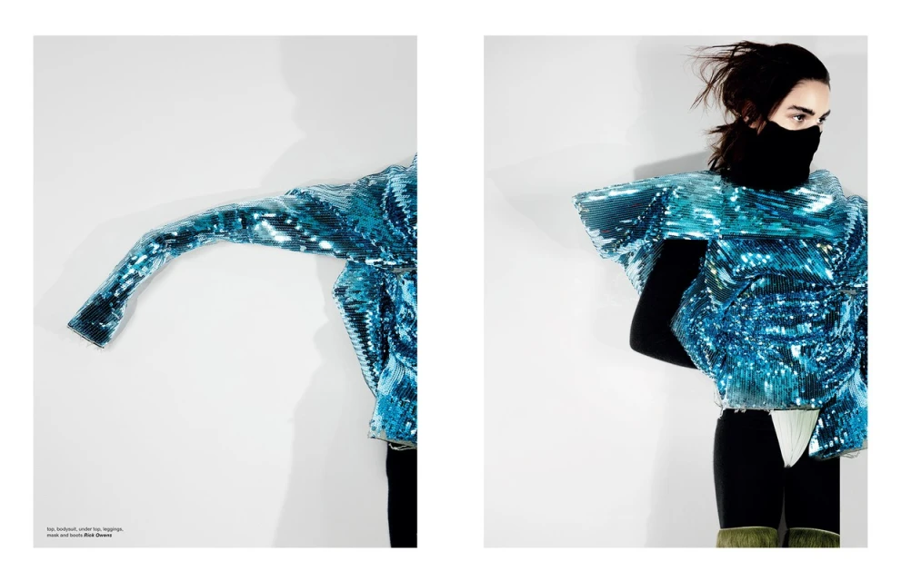 Avant-garde fashion shot of a model wearing a blue, textured jacket, black leggings and a black balaclava against a white backdrop.