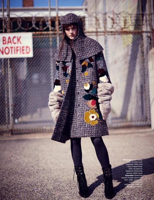Editorial fashion shot of a woman wearing a tweed coat and hat with black tights and lace-up boots, posing in front of a chain-link fence.
