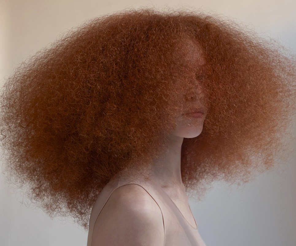Profile portrait of a young adult woman with voluminous red afro hairstyle against a neutral background in a beauty and fashion context.