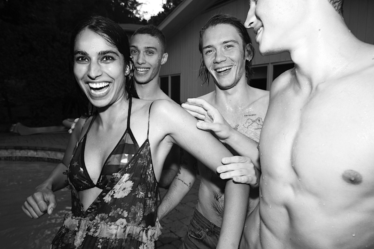 Four young adults are laughing and posing near a swimming pool at night, wearing casual summer clothing and swimsuits, in a social setting.