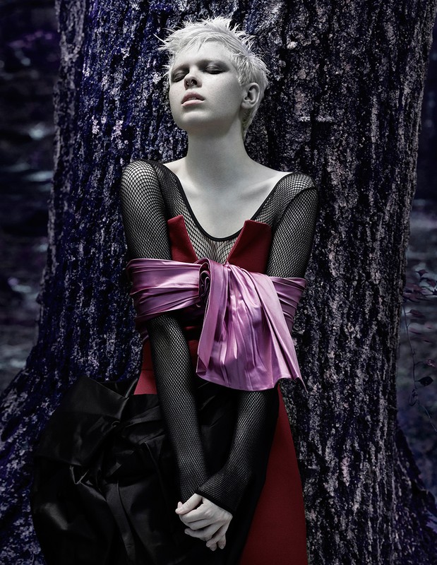 Woman with short blond hair in dark dress stands near large tree in a forest. Red and pink sash tied around waist. Moody scene.