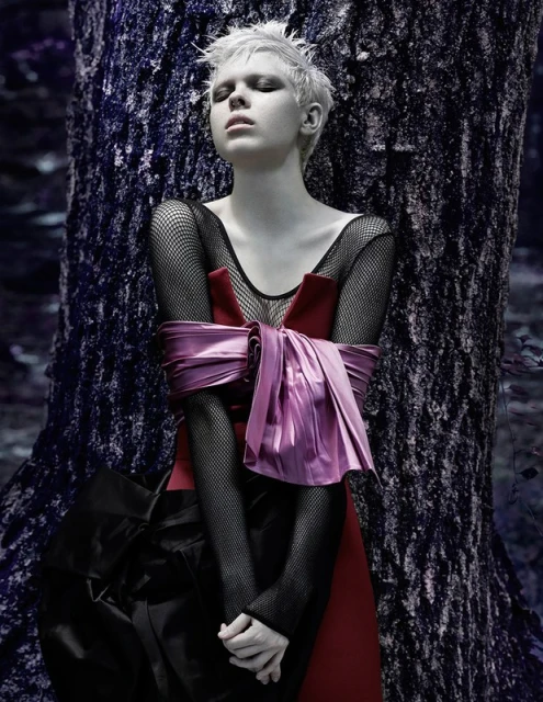 Woman with short blond hair in dark dress stands near large tree in a forest. Red and pink sash tied around waist. Moody scene.