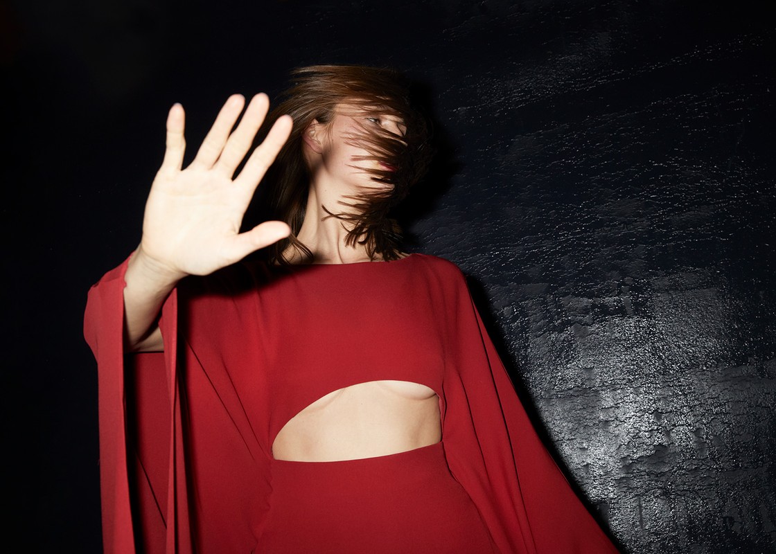 Woman in red dress with cut-out detail posing with hand up in a dark setting, her hair blowing across her face creating motion.