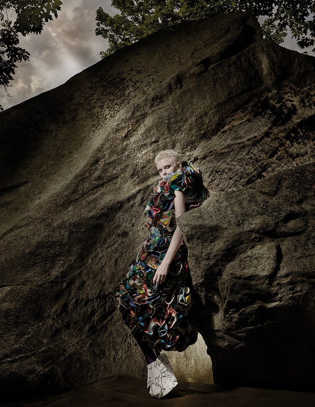A woman with a short haircut and a colorful, sculptural dress poses against a large rock formation in a fashion editorial image.
