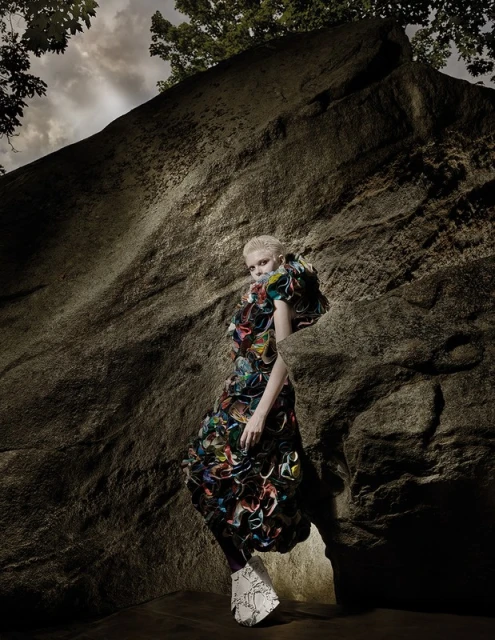 A woman with a short haircut and a colorful, sculptural dress poses against a large rock formation in a fashion editorial image.