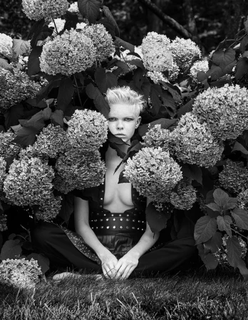 Black and white portrait of a young person sitting amidst blooming hydrangeas, gazing directly at the camera with a serious expression.