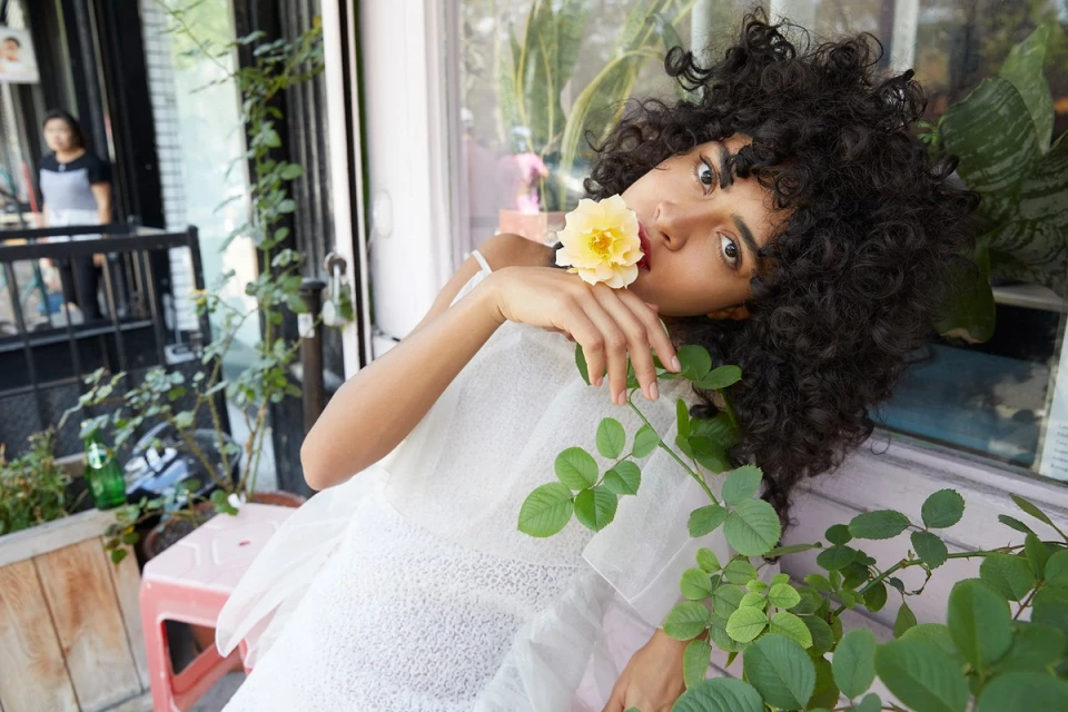 Adult female fashion model with curly hair poses in a white dress with a yellow flower in her mouth, outside on a sunny day.