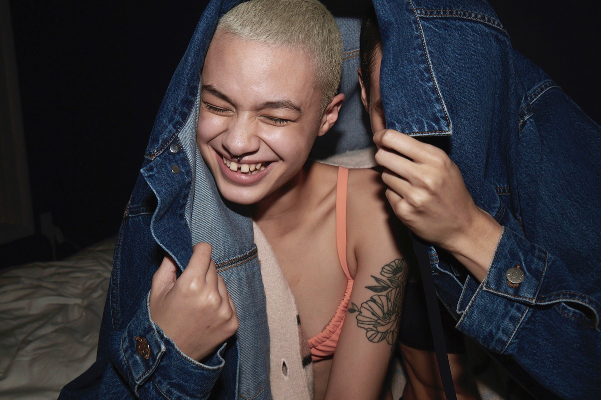 Two people smile under a blue denim jacket indoors. One with fair skin and short blond hair wearing peach-colored bra shows a floral tattoo.