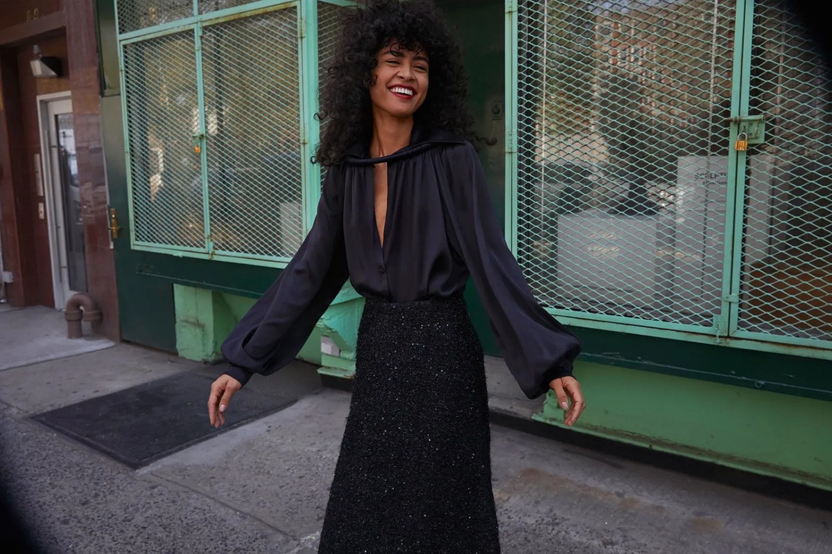 Smiling woman with dark curly hair wearing a dark silk blouse and textured skirt on a city sidewalk in Los Angeles