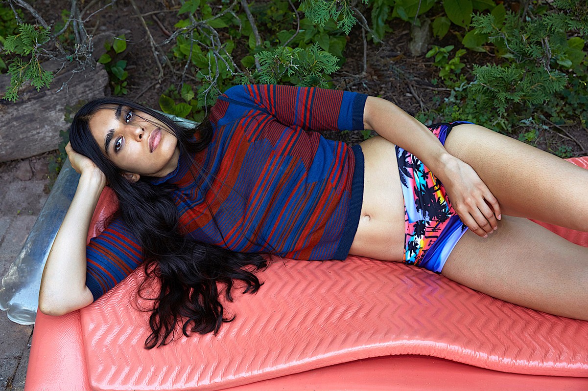 Young woman with long dark hair poses reclining on a coral surface wearing patterned top and swim bottoms outdoors in a leafy setting.
