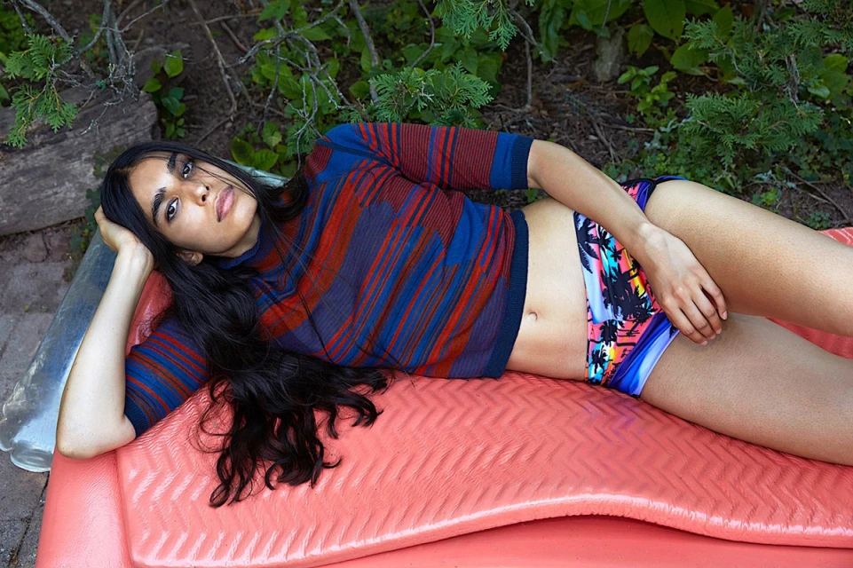 Young woman with long dark hair poses reclining on a coral surface wearing patterned top and swim bottoms outdoors in a leafy setting.