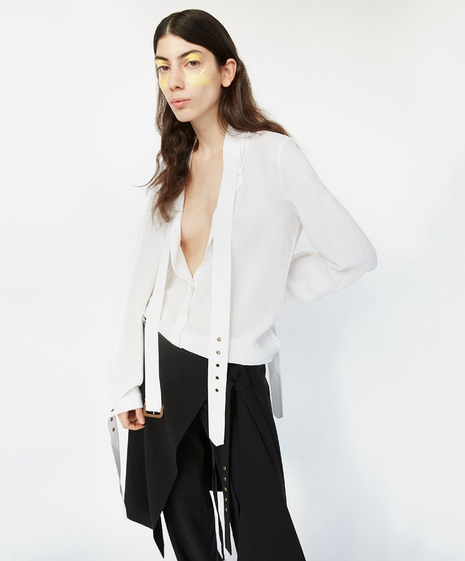 Brunette woman in a white top and black skirt poses against a white background, showcasing makeup and fashion in a studio setting.