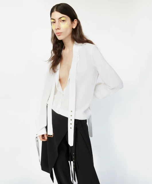 Brunette woman in a white top and black skirt poses against a white background, showcasing makeup and fashion in a studio setting.